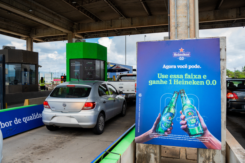 Um carro parado em um pedágio de autoatendimento, ao lado uma placa divulgando a ativação feita pela Onzex.